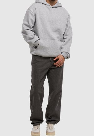 Mann trägt hellgrauen Hoodie, dunkelgraue Jeans in Relaxed-Fit und weiße Sneakers, steht mit einer Hand in der Tasche vor einfachem Hintergrund.