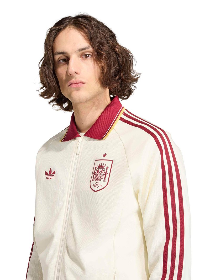 Jeune personne aux cheveux bruns mi-longs portant une veste de sport zippée blanche et rouge avec l'emblème national de l'Espagne.