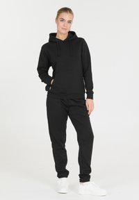 Nainen seisoo yllään musta huppari ja mustat collegehousut valkoisten lenkkareiden kanssa, poseeraten yksinkertaista valkoista taustaa vasten.