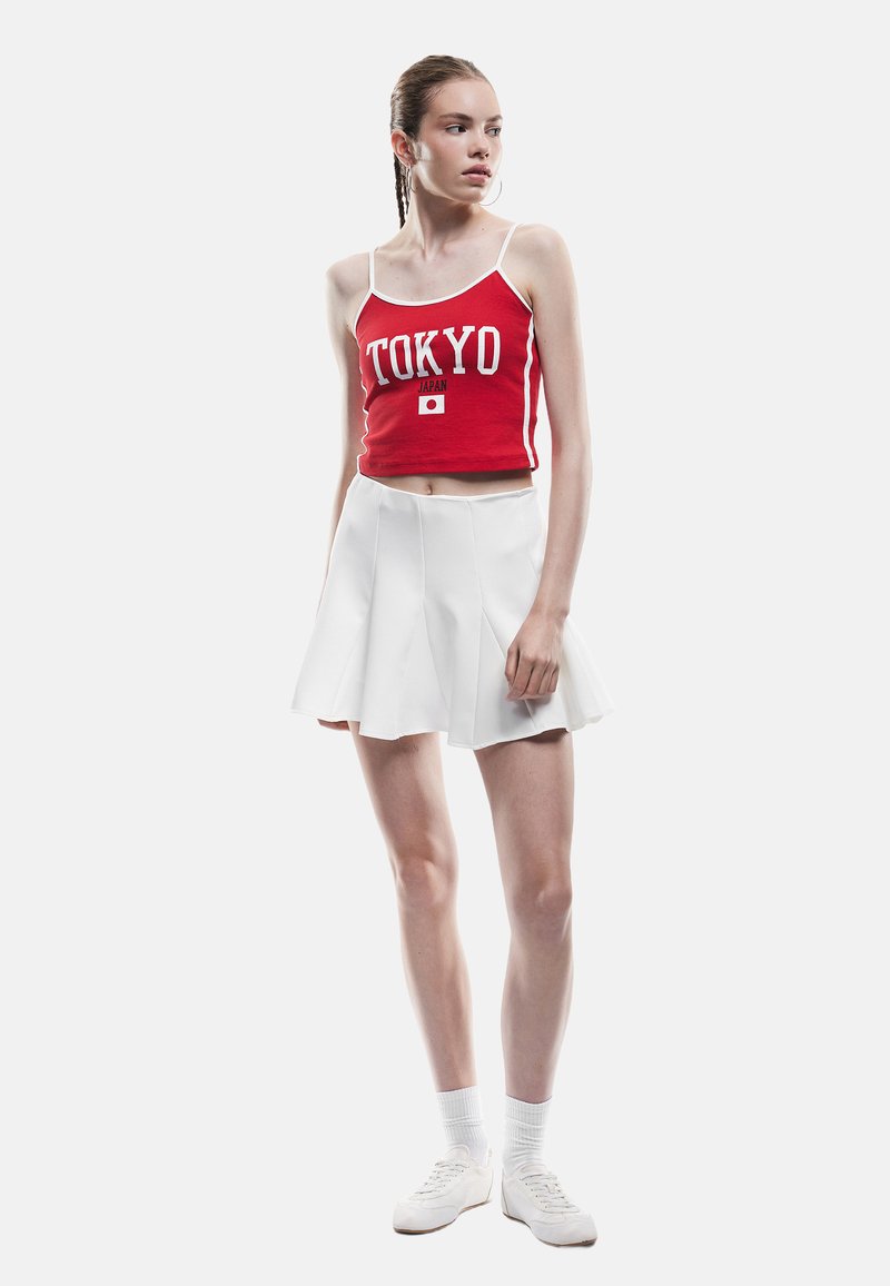 Czerwony krótki top z logo "TOKYO" i grafiką japońskiej flagi, zestawiony z białą mini spódnicą w kształcie litery A. Modelka nosi białe trampki i skarpetki za kostkę.