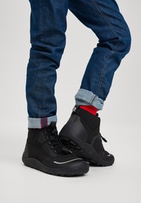 Stivaletti neri alla caviglia con superficie texture, punta rotonda e lacci davanti. Indossati con denim blu arrotolato e calze con accenti rossi. Suole in gomma.
