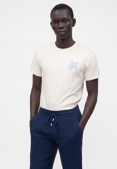 Homme portant un t-shirt beige clair à manches courtes avec un petit motif de tortue bleue et un pantalon bleu marine à cordon, debout les mains dans les poches.