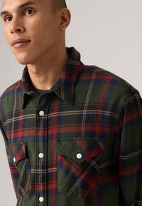 Plaid flannel shirt in green, red, and navy, featuring two chest pockets, white buttons, and a fold-down collar. Textured fabric visible.