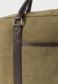 Lærredstaske i olivengrøn med mørkebrune læderdetaljer. Har et robust håndtag og en tekstureret overflade med en lomme foran.