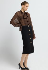 Braune durchsichtige Bluse mit Ripsband-Detail, kombiniert mit einem schwarzen Bleistiftrock mit goldenen Knopfakzenten. Schwarze High Heels runden das Outfit ab.