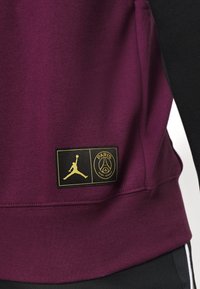 Sweatshirt bordeaux avec une finition texturée, arborant un logo Jordan doré et un badge noir du Paris Saint-Germain sur le côté inférieur.