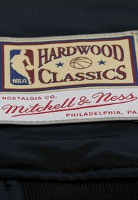 Das gewebte Etikett aus Stoff hat einen goldenen Hintergrund mit rotem und blauem Text; "HARDWOOD CLASSICS" und das NBA-Logo mit einer Basketballgrafik.