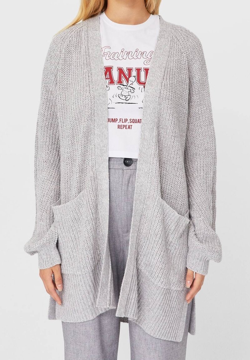 Femme portant un cardigan en maille gris clair avec deux poches avant, par-dessus un t-shirt blanc à motif et un pantalon gris.