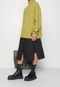Sweatshirt oversize verde oliva, gonna nera a ginocchio con spacchi laterali e bottoni a pressione, abbinata a stivali platform neri lucidi con lacci.