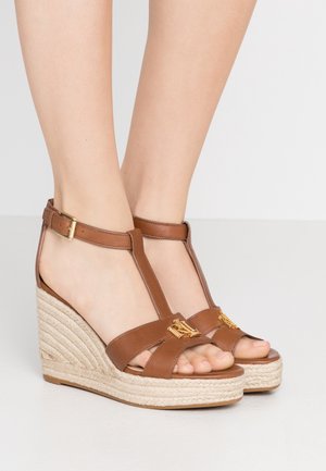 Brune læder kilehæls-sandaler med ankelremme og guldfarvet logodetalje, båret på bare fødder mod en neutral baggrund.