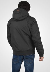Sort hættetrøje med en glat tekstur, elastiske manchetter og en tætsiddende talje, båret over blå denimjeans med synlige syninger og lommer.