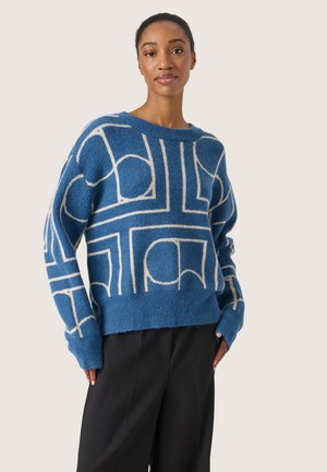 Blå strikket sweater med et geometrisk mønster i creme, rund halsudskæring og ribbede kanter, samt sorte skræddersyede bukser.