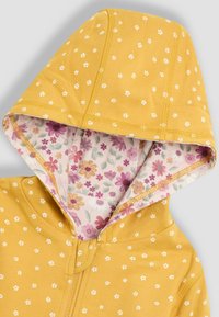 Giacca gialla con cappuccio, decorata con piccoli motivi floreali bianchi e una fodera a stampa floreale all'interno del cappuccio. Il materiale sembra morbido e tessurizzato.