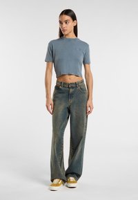 T-shirt blu cropped con maniche corte, abbinata a jeans blu chiaro larghi e sneakers gialle. Il denim presenta una lieve texture e cuciture a contrasto.
