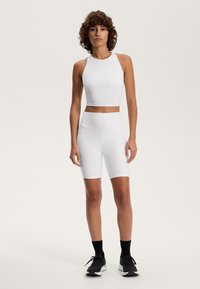 Bijeli sportski crop top i odgovarajuće kratke biciklističke hlače visokog struka, izrađeni od glačane, rastezljive tkanine, nosi se s crnim trkačkim cipelama.