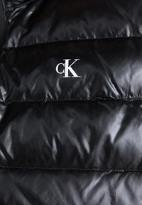 Crna puffer jakna s kocedim dizajnom, sjajnom teksturom i bijelim "cK" logotipom na gornjoj lijevoj strani.