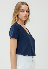 Granatowa koszulka z dekoltem w serek, wykonana z miękkiego materiału, z krótkimi rękawami i luźnym krojem. Ozdobiona dyskretnymi przeszyciami.