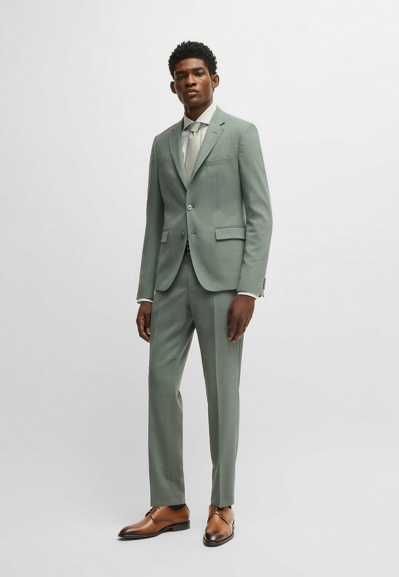 Costume vert clair au design ajusté, pantalon à devant plat et blazer à un bouton. Assorti à une chemise à rayures claires et des chaussures brunes.