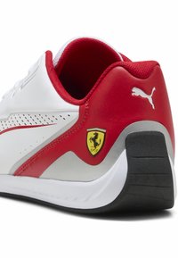Sneakers bianchi e rossi con pannelli laterali perforati, dettagli in argento, suola in gomma nera e un emblema giallo della Ferrari sul tallone.