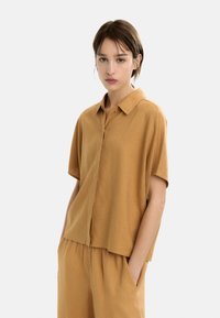 Młoda kobieta z krótkimi brązowymi włosami, ubrana w luźną beżową koszulę z krótkim rękawem i pasujące spodnie, stojąca z rękami w kieszeniach na białym tle.