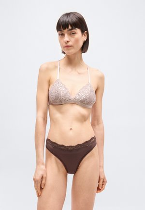 Γυναίκα με κοντά σκοτεινά μαλλιά που φοράει ροζ ανοιχτό δαντελένιο μπραλέτα και σκούρο καφέ εσώρουχο με δαντέλα, στέκεται μπροστά σε απλό φωτεινό φόντο.