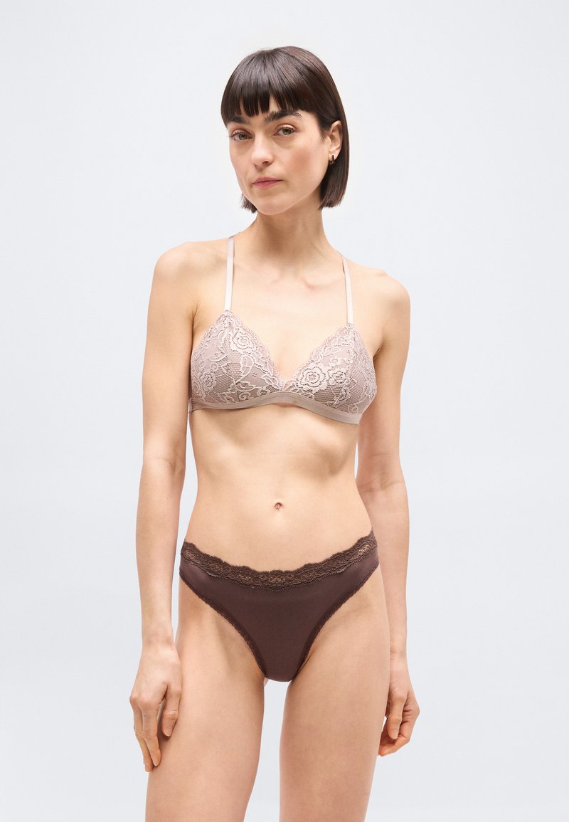 Γυναίκα με κοντά σκοτεινά μαλλιά που φοράει ροζ ανοιχτό δαντελένιο μπραλέτα και σκούρο καφέ εσώρουχο με δαντέλα, στέκεται μπροστά σε απλό φωτεινό φόντο.