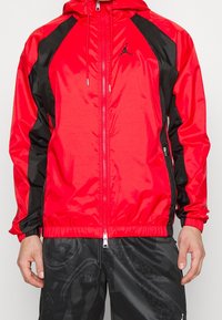 Veste coupe-vent rouge et noire avec une finition brillante, capuche, fermeture éclair et détail logo sur la poitrine ; poignets et ourlet élastiques.