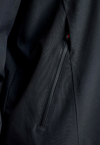 Giacca nera in tessuto con tasca con zip nascosta. Il materiale ha una consistenza liscia, con dettagli di cucitura lungo le cuciture.
