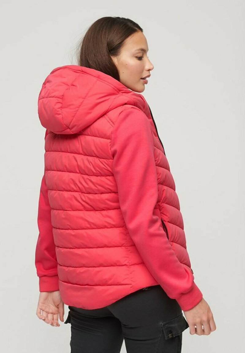 Rote gepolsterte Weste mit Kapuze, mit horizontalen Nähten, Reißverschluss und elastischen Bündchen, getragen über einem Langarmoberteil.