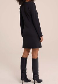 Czarna sukienka z długim rękawem o dopasowanej sylwetce, sięgająca do kolan, z gładką fakturą, zestawiona z czarnymi, wysokimi butami na obcasie.