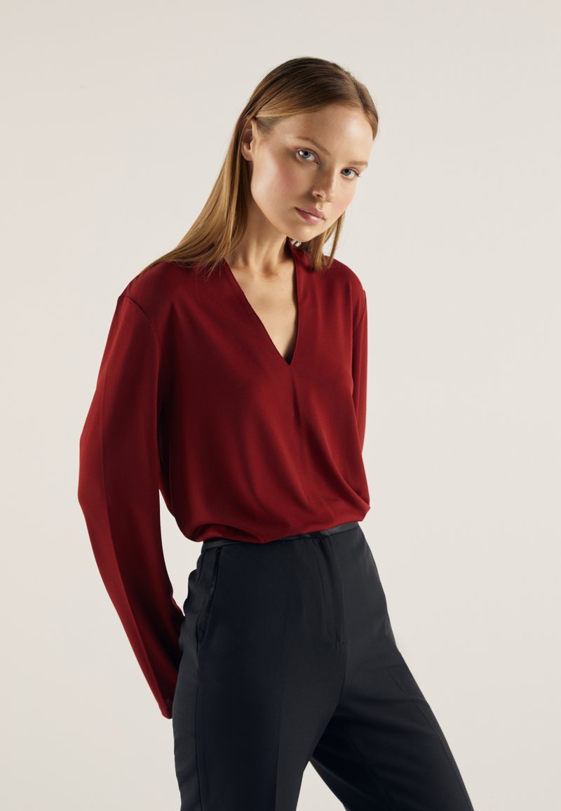 Bluza roșie cu decolteu în V și mâneci lungi, din material neted, asortată cu pantaloni negri mulați, evidențiind un design minimalist și linii fine.