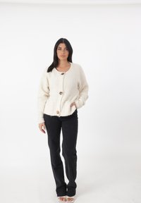 Cardigan bianco e peloso con tre grandi bottoni e due tasche frontali, abbinato a pantaloni neri e scarpe aperte su uno sfondo chiaro.