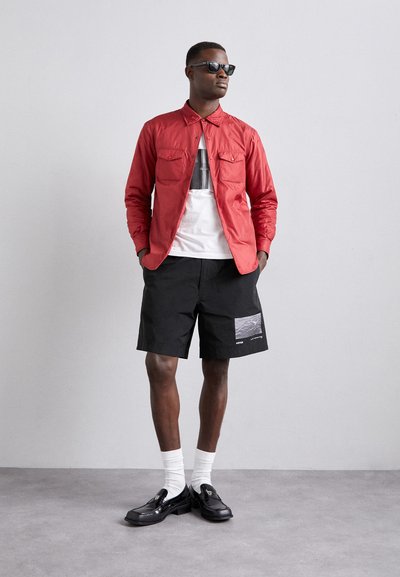 Veste légère rouge avec des fermetures à boutons, shorts noirs avec un patch graphique, chaussettes blanches et mocassins noirs. Lunettes de soleil noires portées.