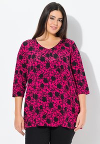 Rosa und schwarze florale Tunika mit Dreiviertelärmeln, V-Ausschnitt, lockerer Passform und strukturierter Stoff. Darunter werden schwarze, einfarbige Hosen getragen.