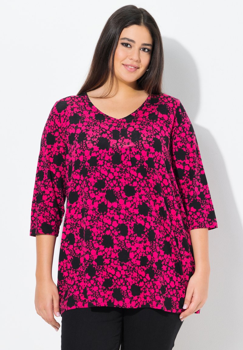 Rosa und schwarze florale Tunika mit Dreiviertelärmeln, V-Ausschnitt, lockerer Passform und strukturierter Stoff. Darunter werden schwarze, einfarbige Hosen getragen.