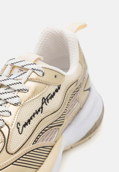 Beige sneakers i mesh och lädermaterial, med svarta accenter, en texturerad sida och en vit gummisula. Signaturdetalj på sidan.