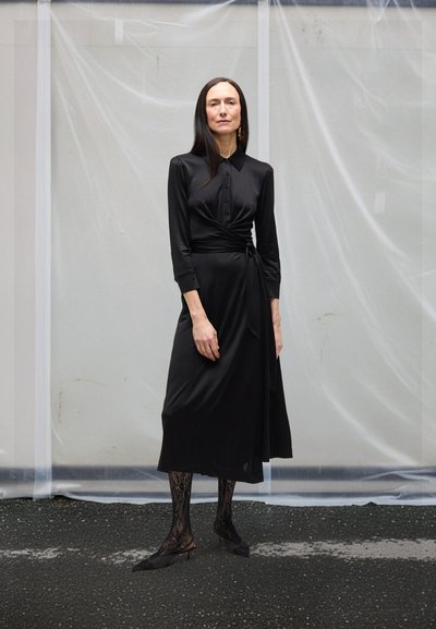 Ženska z dolgimi temnimi lasmi, oblečena v črno dolgoročno obleko z zavitkom, črne čipkaste nogavice in črne koničaste čevlje, stoji na asfaltu.