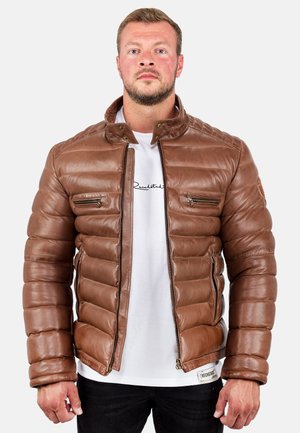 Mann, der nach vorne blickt, trägt eine braune gesteppte Lederjacke über einem weißen T-Shirt und schwarzen Hosen vor einem schlichten Hintergrund.