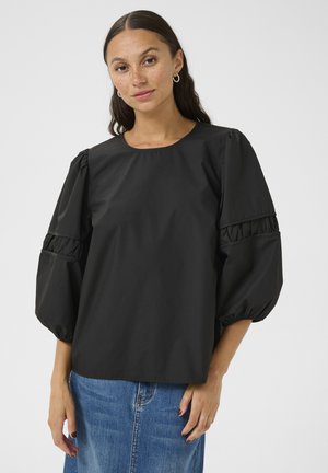 Blouse noire tissée avec un col rond, des manches bouffantes et des accents froncés aux poignets. Associée à une jupe en denim bleu.