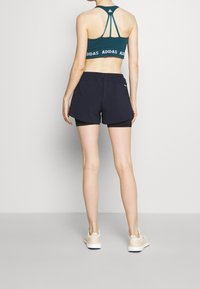 Sujetador deportivo verde azulado con la marca ADIDAS, pantalones cortos negros de entrenamiento sobre mallas negras ajustadas, y zapatillas ligeras con material texturizado.