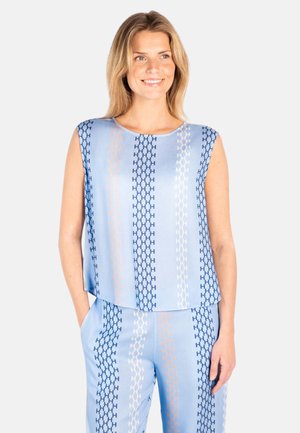 Vrouw draagt lichtblauw mouwloos topje en bijpassende broek met verticale, meerkleurige kettingschakelpatronen, glimlacht en staat met handen in de zakken.