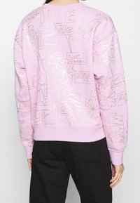 Rosa sweatshirt med en mängd olika Nike-logotyper, blommönster och text. Har en något oversized passform och ribbade ärmslut och nederkant.