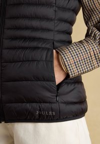 Hand in der Tasche einer schwarzen gesteppter Weste über einem beigen karierten Hemd mit goldenem Armreif, weiße Hose und sichtbares „Joules“-Logo auf der Weste.