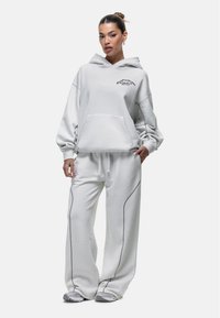 Felpa grigia chiara oversize con tasca frontale, abbinata a pantaloni della tuta coordinati con dettagli di cucitura scura. Entrambi gli articoli hanno una texture morbida.
