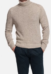 Béžový pletený rolák se žebrovanými manžetami a lemem, s texturovaným vzorem a volným střihem, nošený s tmavě modrými kalhotami.