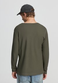 Camicia verde a maniche lunghe in tessuto waffle textured, con scollo ampio e berretto nero indossato al contrario. Vestibilità casual con polsini a coste.