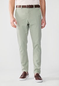 Pantalons chinos vert clair avec une texture lisse et une coupe droite, associés à une ceinture en cuir marron et des mocassins marron foncé.