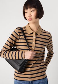 Giovane donna che indossa una polo a maniche lunghe marrone e nera a righe, con una borsa a tracolla in pelle nera, che guarda di lato.