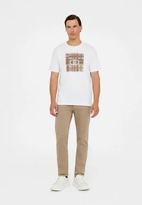 Weißes kurzärmliges T-Shirt mit braunem Karomuster auf der Vorderseite, kombiniert mit beiger Hose und weißen Turnschuhen.