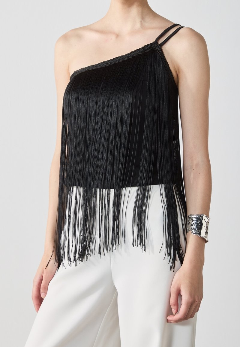 Femme portant un haut noir asymétrique à franges avec des bretelles doubles et un pantalon blanc, accessoirisée d'un bracelet manchette en argent.